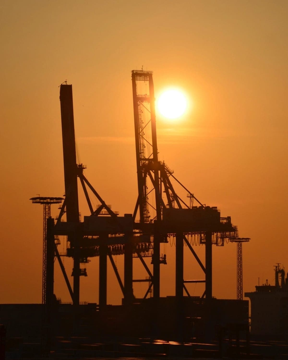 Container cranes standing against the sun low over a port at day's end.
